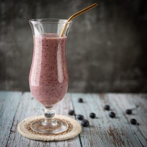A glass with a purple smoothie and a straw on a table with scattered blueberries.