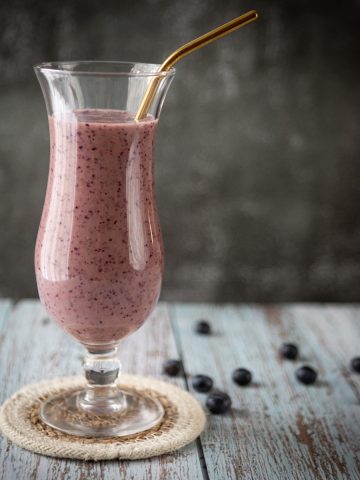 A glass with a purple smoothie and a straw on a table with scattered blueberries.