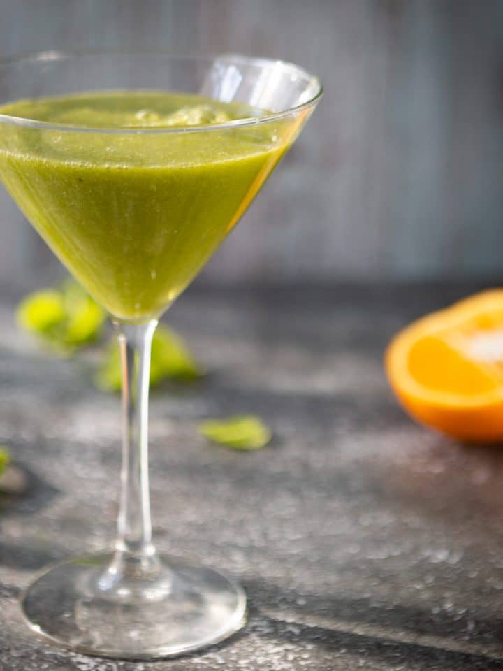 A glass with bright green smoothie, on a grey background, with orange in the background.