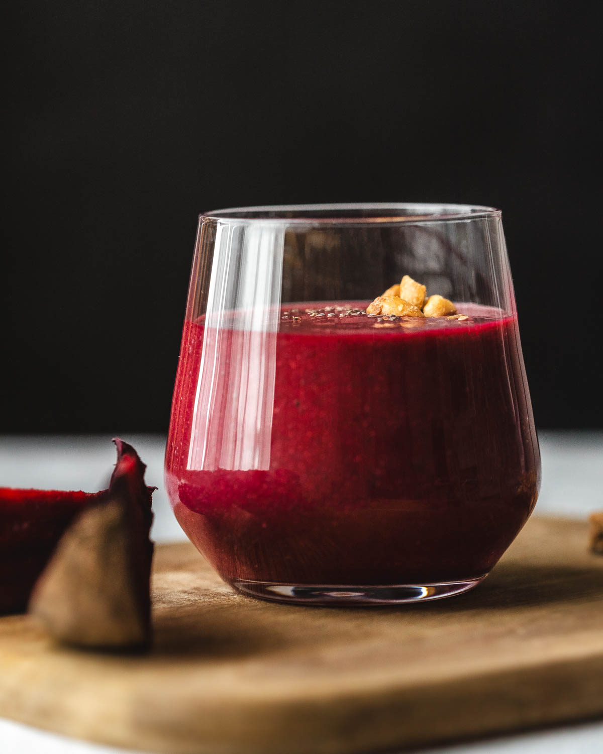 A side view of a glass filled with red beetroot and strawberry oat smoothie, on a wooden cutting board along with a couple of beetroot wedges.