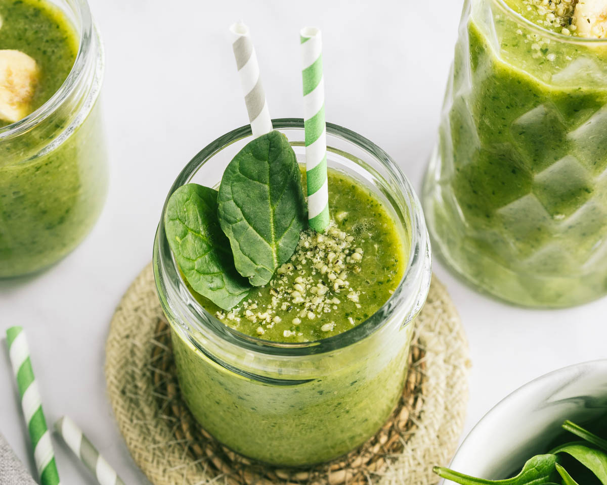 A glass of a green spinach smoothie on a round coaster, with two straws and a couple of spinach leaves in the glass; two other glasses in the background.