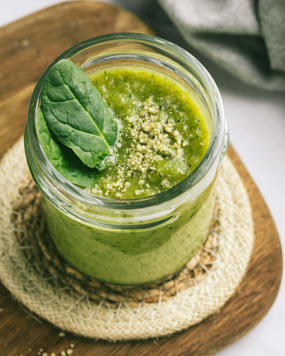 A jar of green spinach smoothie with two spinach leaves in it, on a round coaster on a wooden cutting board.