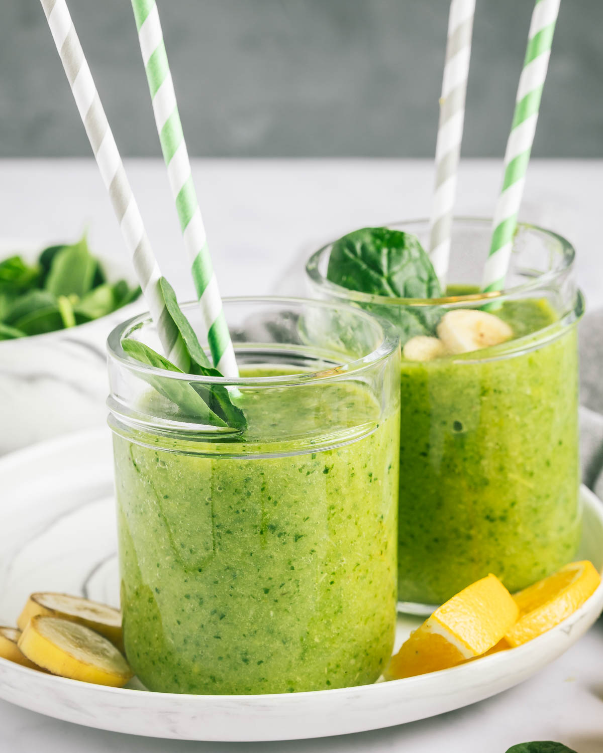 Two jars of green spinach and citrus smoothie on a round plate, with two straws in each jar.