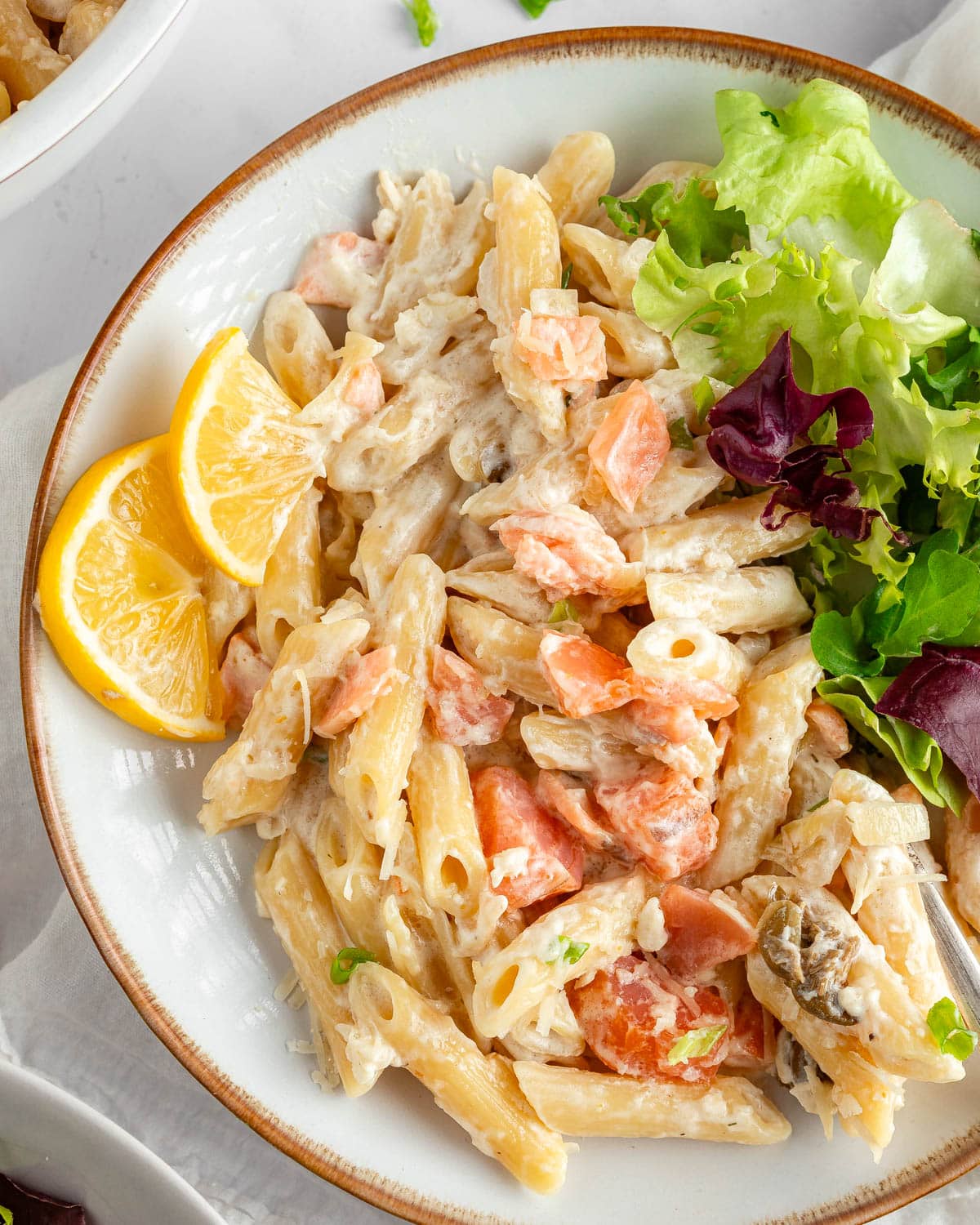 A served smoked salmon pasta salad with lemon wedges and green leaf salad on the side.