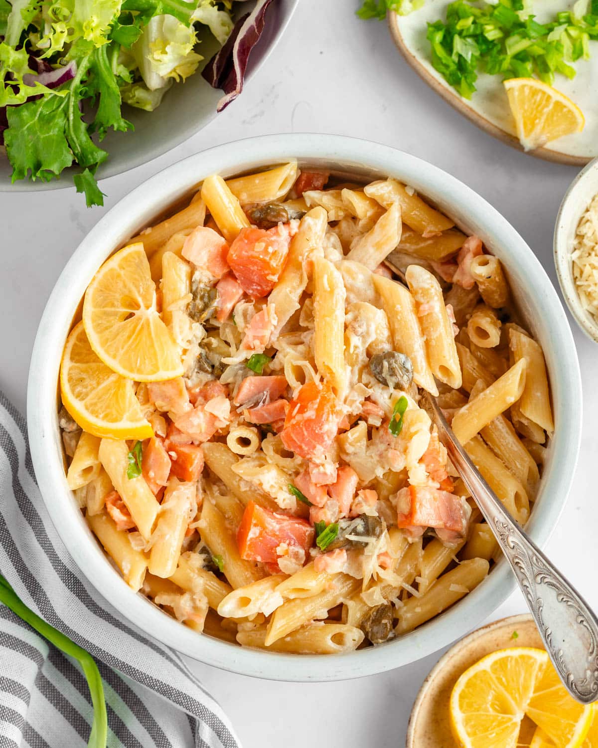 An overhead shot of pasta salad with smoked salmon chunks, on a white table surface with bowls of different ingredients around it.