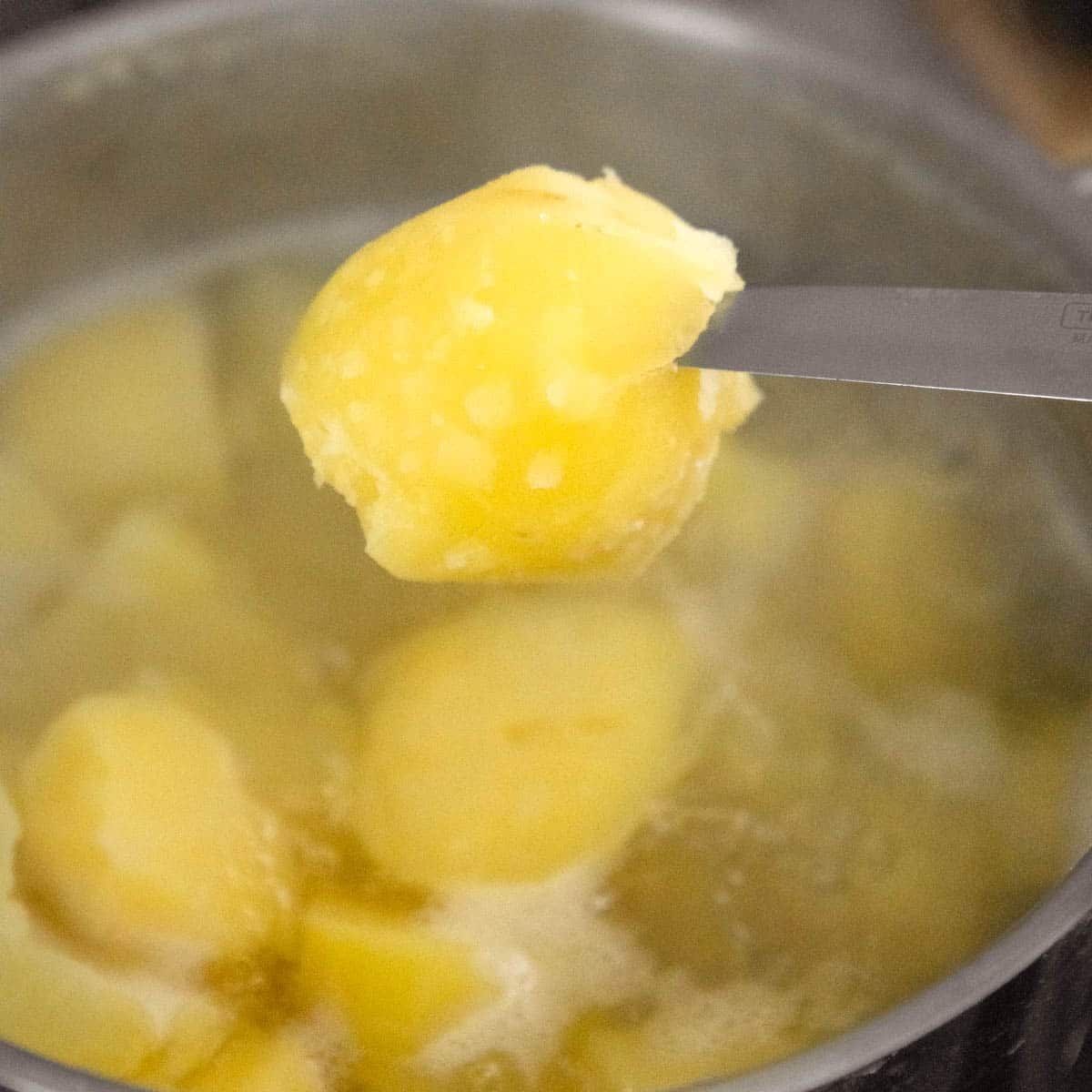 A boiled potatoe on the tip of a knife above a pot with boiling potatoes.