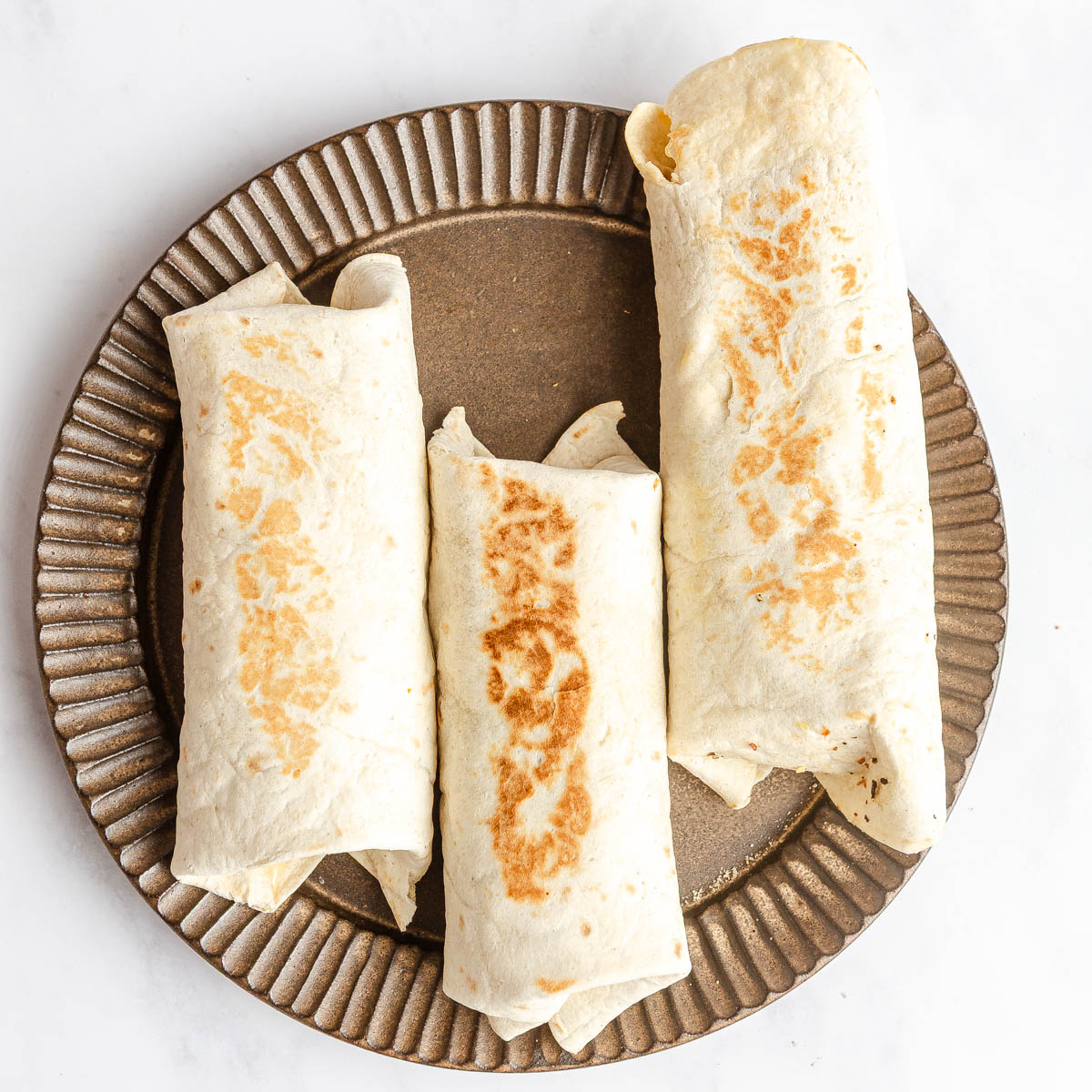 Three closed tortilla wraps on a round dark brown plate.