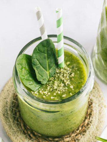 A glass of a green spinach smoothie on a round coaster, with two straws and a couple of spinach leaves in the glass.