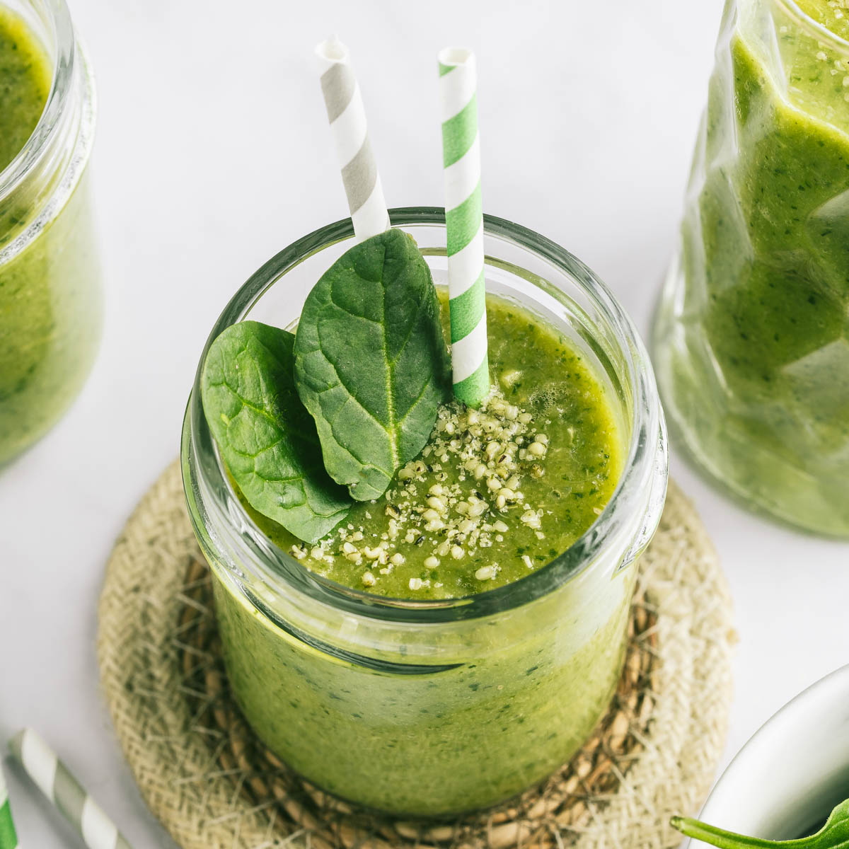 A glass of a green spinach smoothie on a round coaster, with two straws and a couple of spinach leaves in the glass.