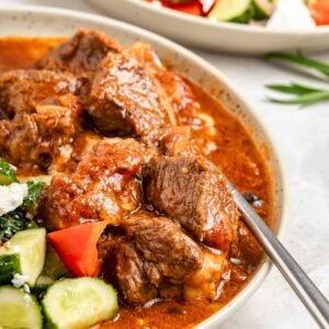 Greek beef stifado served along fresh Greek salad, with a fork submerged.