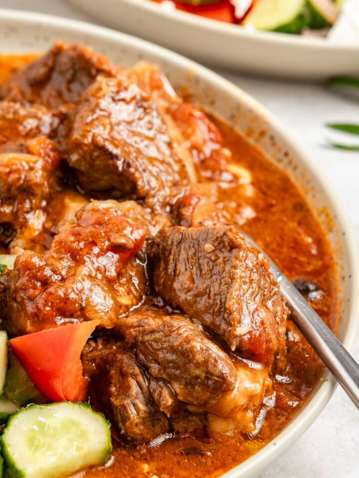 Greek beef stifado served along fresh Greek salad, with a fork submerged.