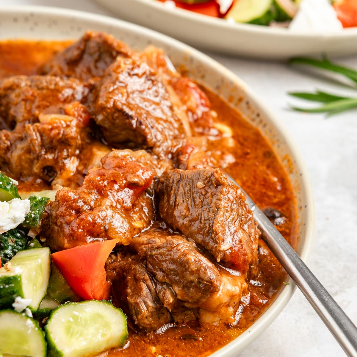Greek beef stifado served along fresh Greek salad, with a fork submerged.