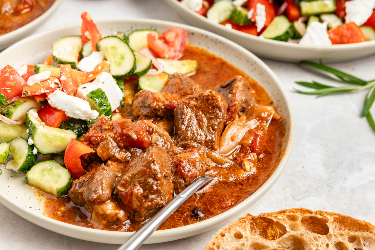 Beef stifado served with greek salad, with a fork submerged in the juicy stifado.