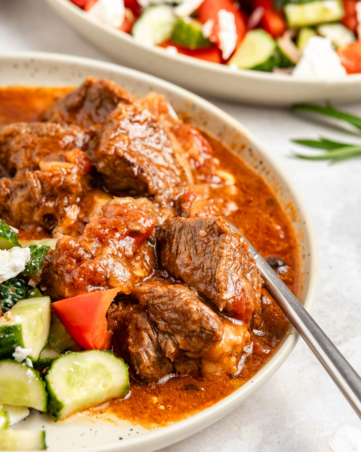 A closeup shot of a plate with greek salad and beef stifado, with another plate in the background.